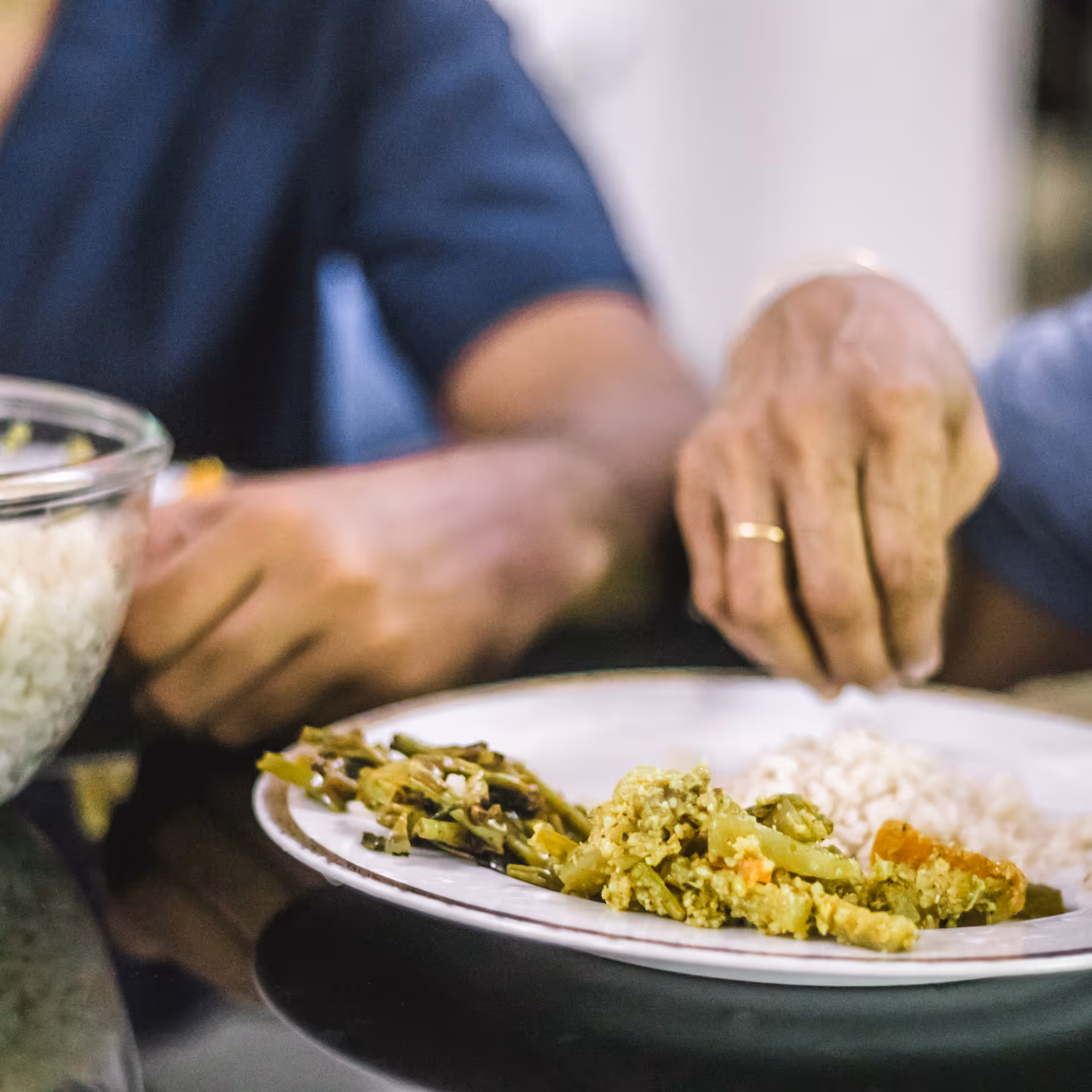 Close-up of two people sitting at a table with a plate of food containing rice and cooked vegetables. One person is wearing a gold ring and is reaching towards the plate.