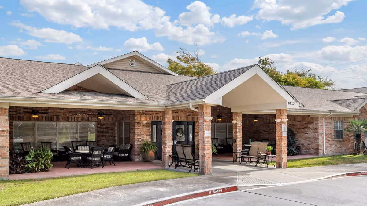 Brick single-story facility front with a covered entrance/porte-cochere, outdoor seating area, and a small lawn under a partly cloudy sky.