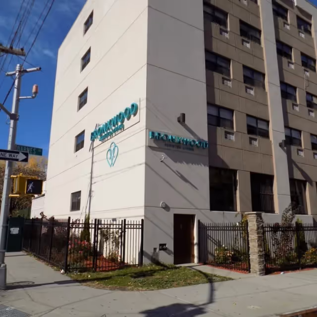 Exterior view of Bronxwood Assisted Living building on a sunny day, showing a multi-story structure with beige and brown walls, a fenced garden area, and signage displaying the facility's name.