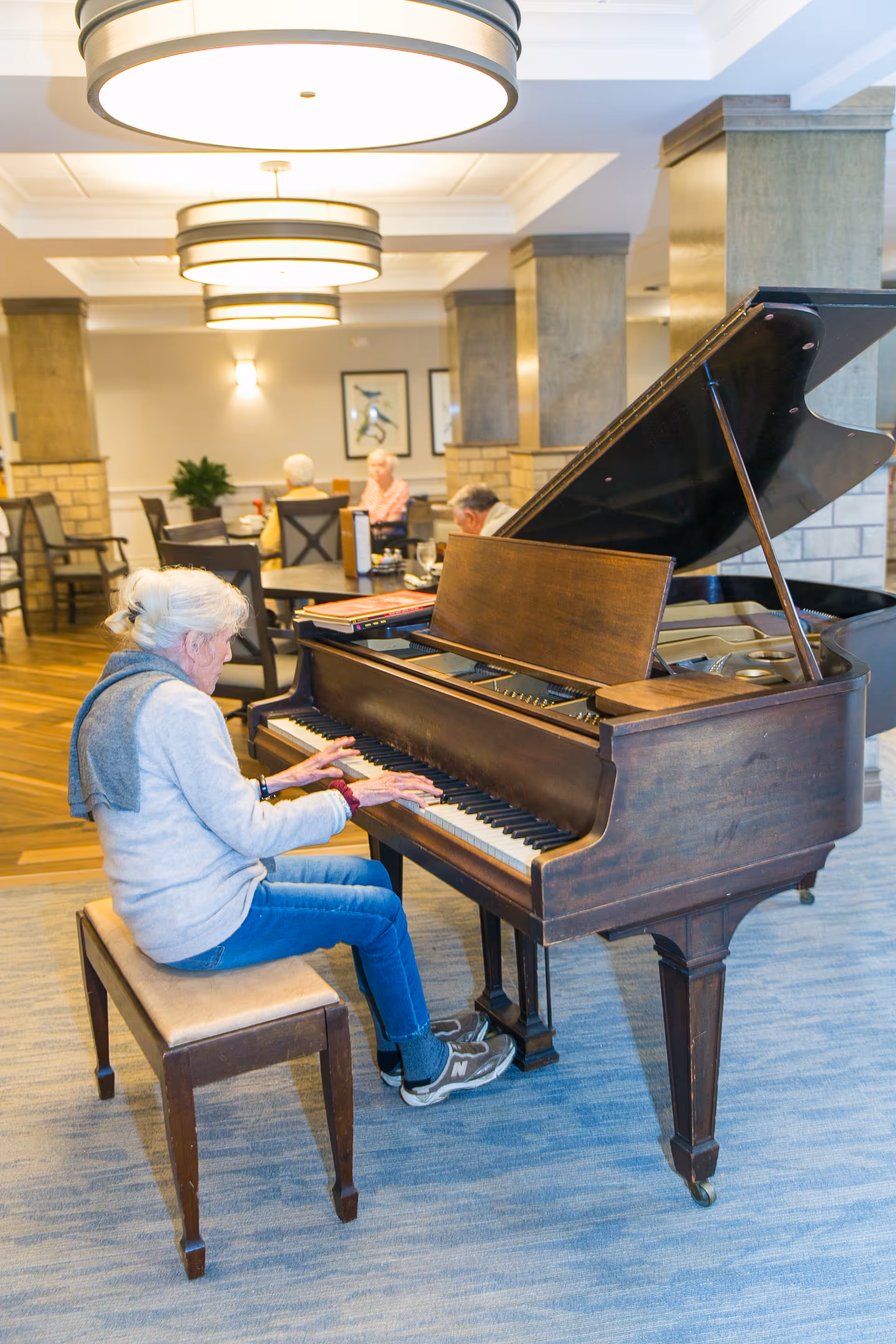 An elderly woman with white hair tied back is playing a grand piano in a well-lit common area. The room has large round ceiling lights, wooden floors, and several tables with other elderly people seated and engaged in conversation. The setting appears cozy and inviting with neutral tones and framed artwork on the walls.