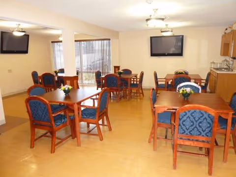 A dining room with several wooden tables and blue cushioned chairs arranged neatly. Each table has a small flower arrangement. There are two wall-mounted flat-screen TVs and large windows with vertical blinds letting in natural light. The room has light-colored walls and a wooden floor.