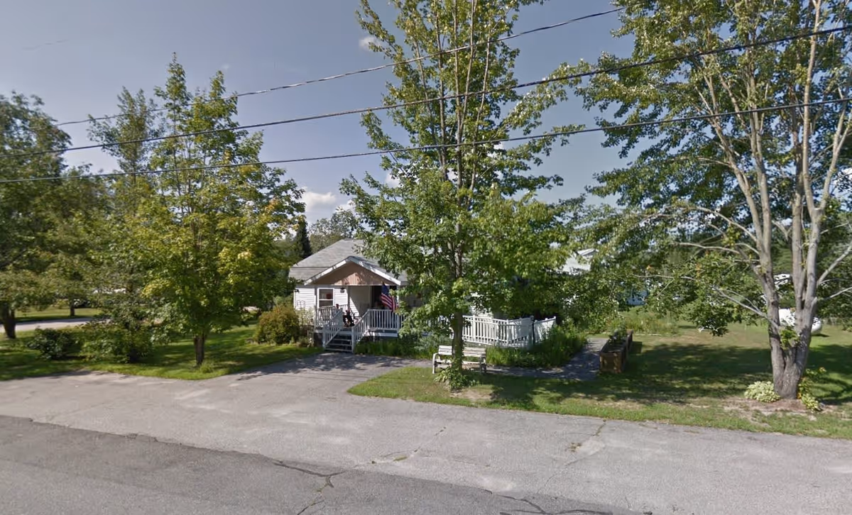 Small single-story house with a front porch, surrounded by green trees and grass under a clear blue sky. There is a paved driveway and a bench near the front yard.