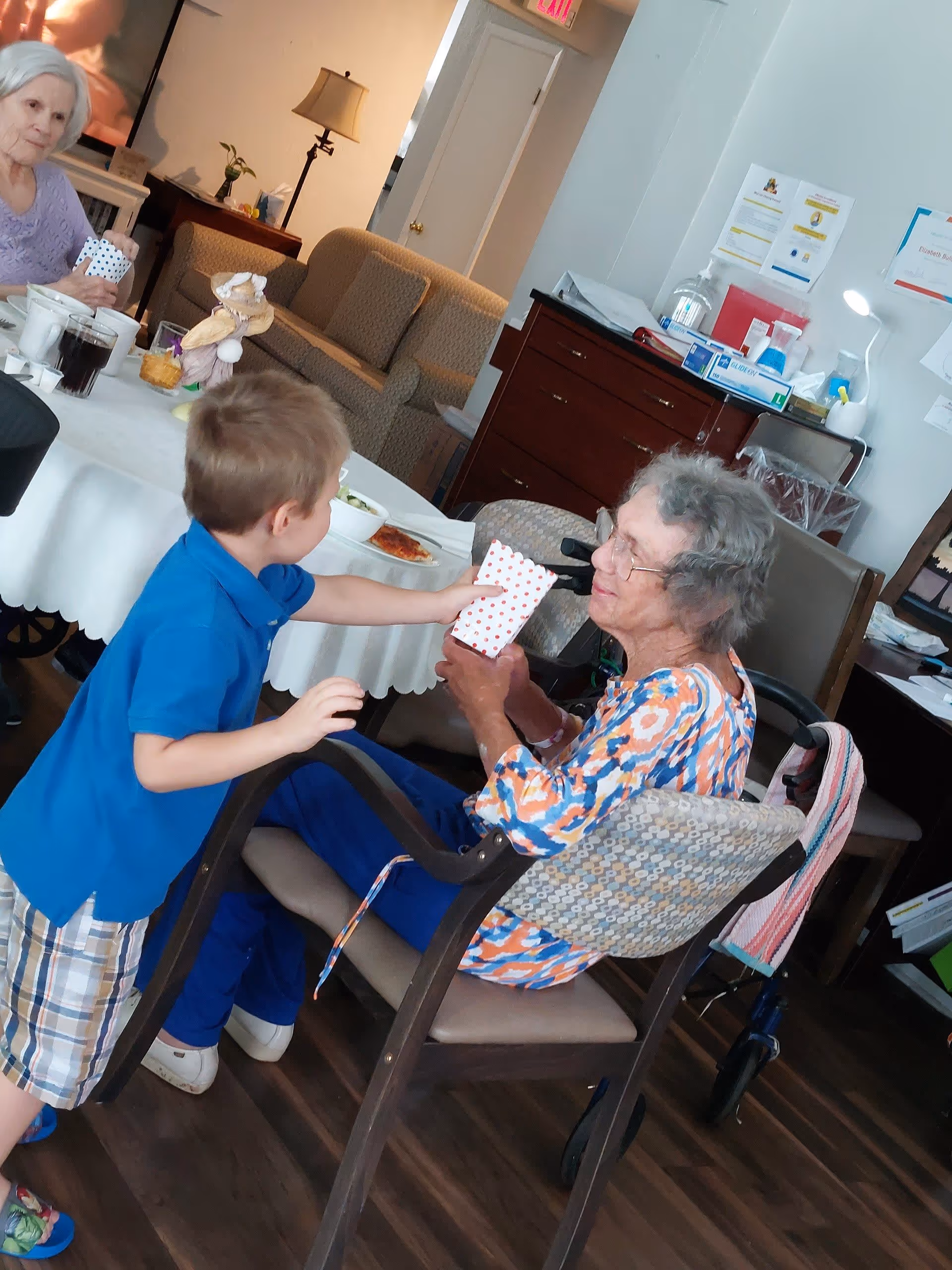 A young boy in a blue shirt and plaid shorts is handing a polka-dotted paper bag to an elderly woman with gray hair and glasses, who is seated in a wheelchair. Another elderly woman with white hair is sitting at a table nearby, holding a similar polka-dotted bag. The room has a cozy living area with a couch, a lamp, and a wooden cabinet with various items on top.