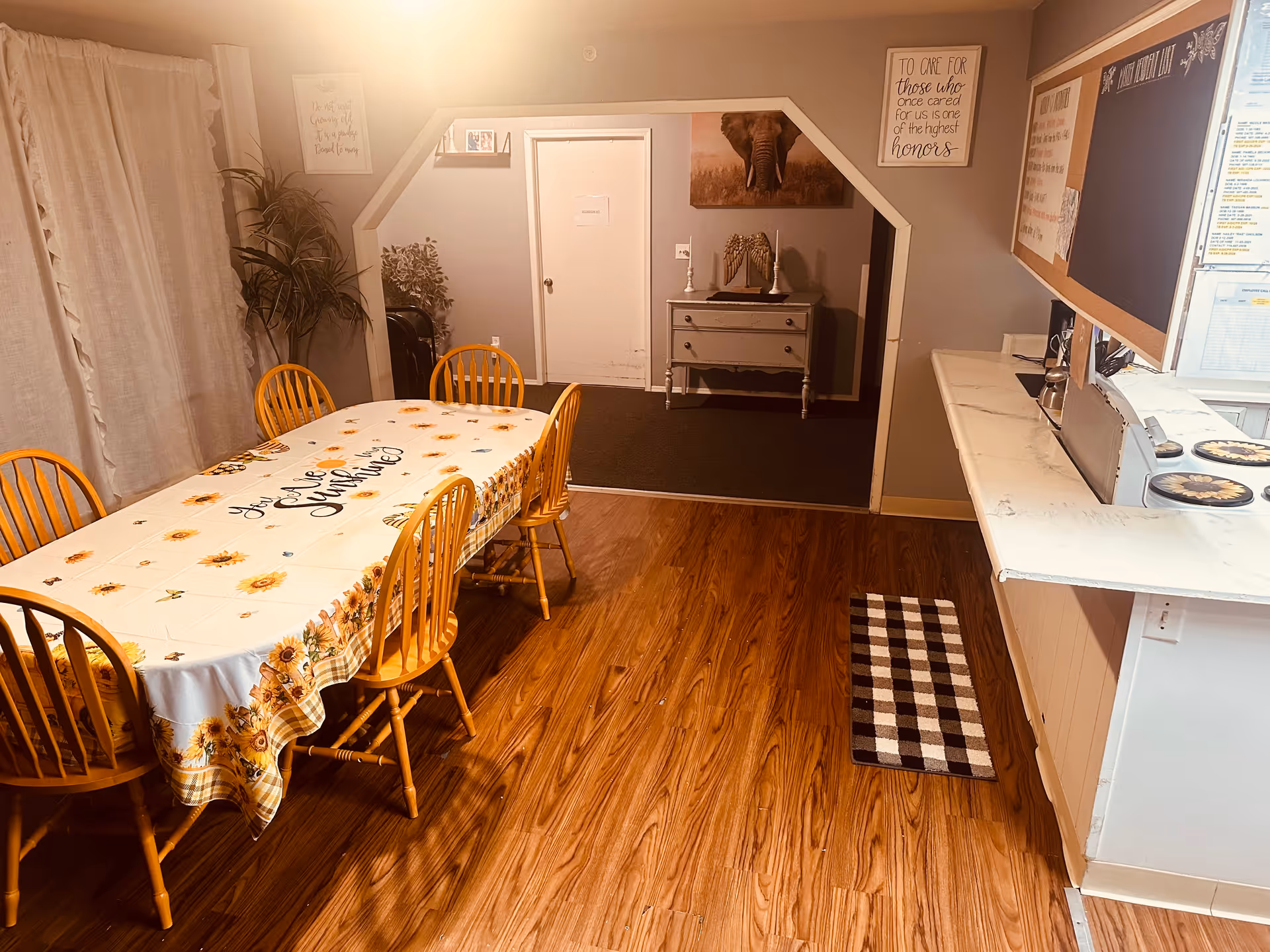 A dining area with a wooden table covered by a sunflower-themed tablecloth and six wooden chairs. The room has wood flooring, a white curtain on the left, and a small cabinet with decorative items against the far wall. There are framed pictures and quotes on the walls, and a counter with a checkered rug in front of it on the right side.