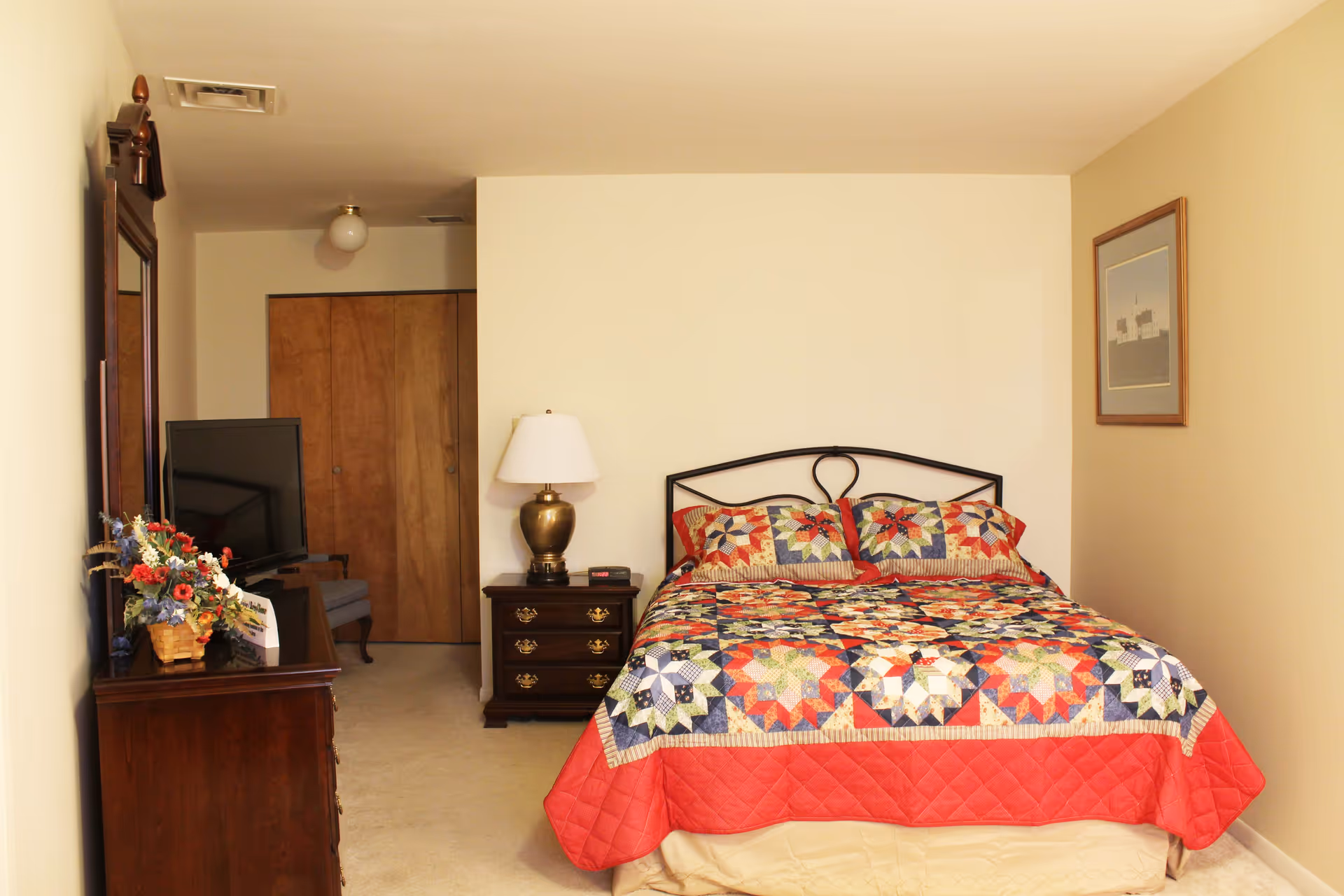 Bedroom with a double bed covered by a colorful patchwork quilt, nightstand with lamp, dresser with TV and floral arrangement, and closed closet doors.