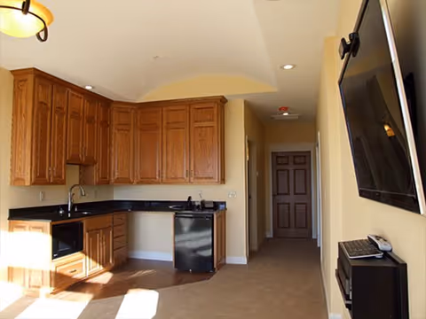 Interior view of a kitchen area with wooden cabinets, a black countertop, a sink, a microwave, and a small black refrigerator. The room has beige walls and carpeted flooring extending into a hallway with a closed door at the end. A flat-screen TV is mounted on the wall to the right.