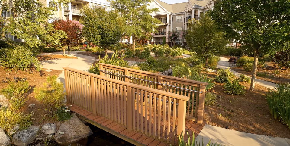 A wooden footbridge over a small stream in a landscaped garden area with trees, shrubs, and walking paths. In the background, there are multi-story residential buildings with balconies surrounded by greenery.