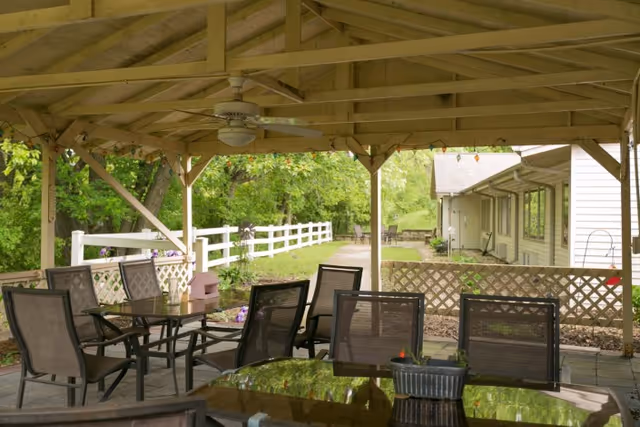 Covered outdoor patio area with a wooden roof structure, ceiling fan, and several glass-top tables surrounded by mesh chairs. The patio overlooks a garden area with a white fence and green trees in the background.