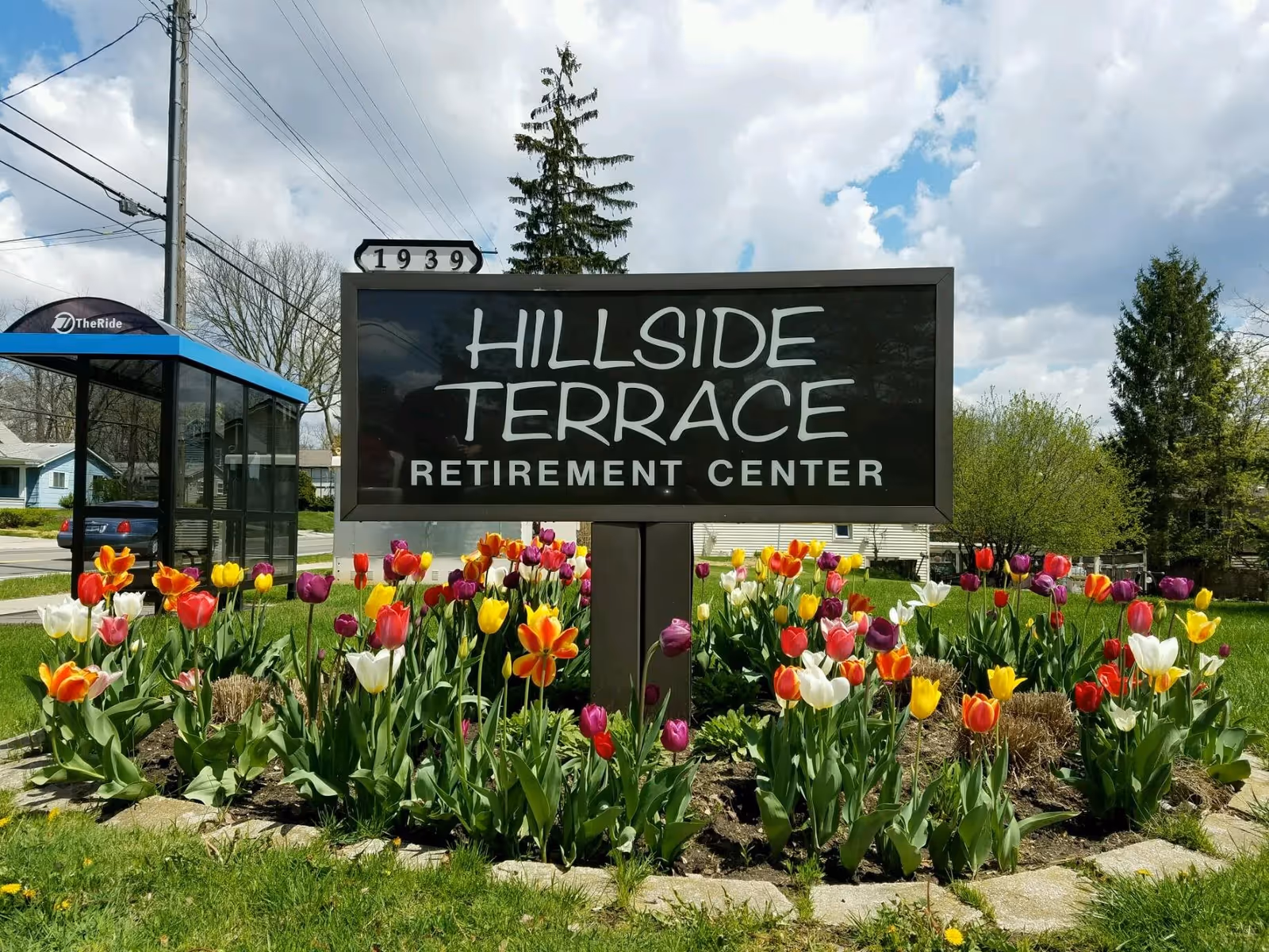 A roadside sign reading "HILLSIDE TERRACE RETIREMENT CENTER" surrounded by colorful tulips under a partly cloudy sky.