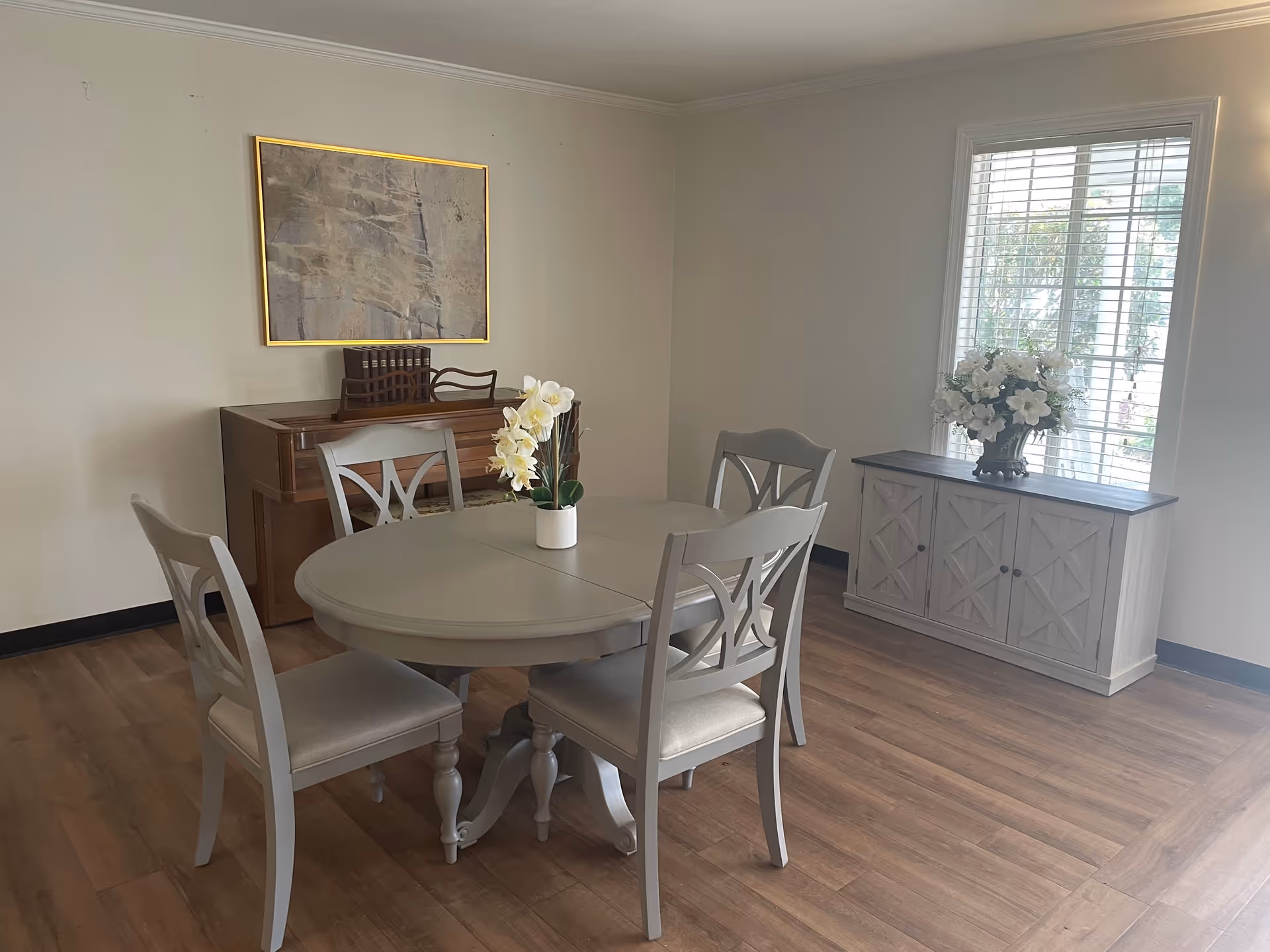 A dining area with a round gray table and four matching chairs. A vase with white flowers is placed on the table. In the background, there is a wooden piano with a set of books on top and a framed abstract painting hanging above it. To the right, a window with white blinds allows natural light into the room, and below the window is a light-colored cabinet with a floral arrangement on top. The floor is wooden, and the walls are painted light beige.