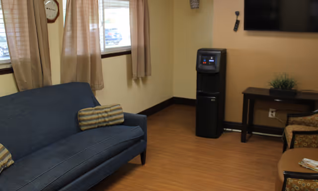 A small sitting area with a blue couch with striped pillows, two windows with beige curtains, a water cooler, a wall-mounted TV, a small table with a plant, and two patterned armchairs around a round table with magazines.