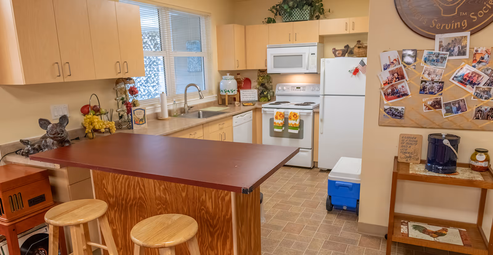 Communal kitchen with a central island and two stools, cabinets, sink, stove, refrigerator, and a photo-covered bulletin board.