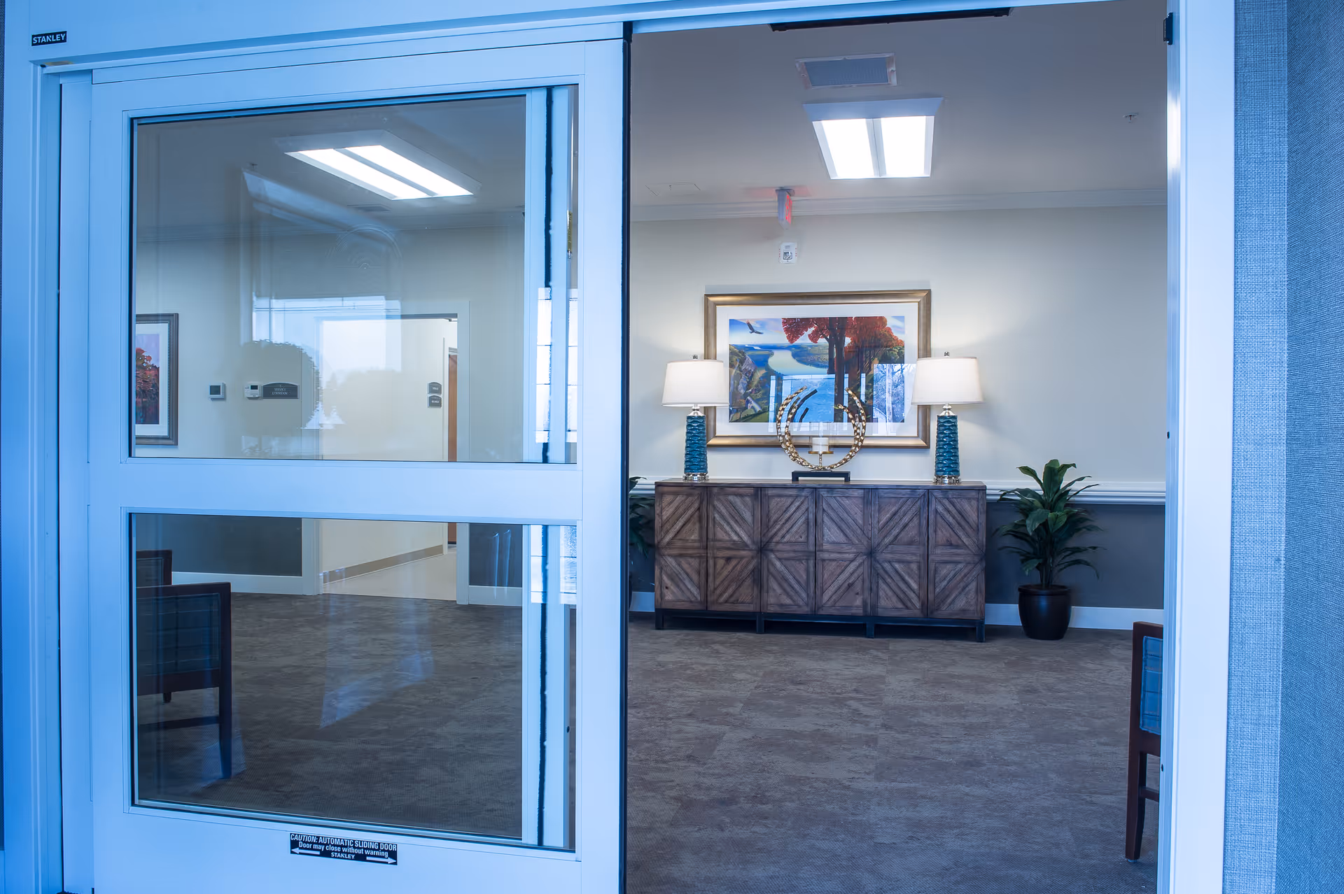 View through automatic sliding glass doors into a reception area with a wooden console table, two lamps, artwork, and potted plants.