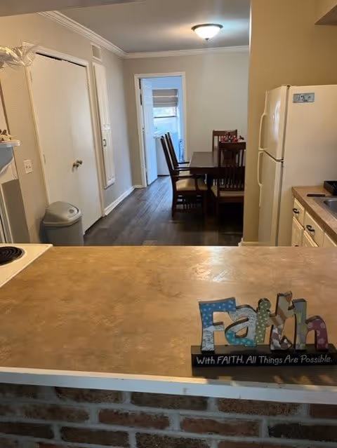 View of a kitchen counter with a decorative sign that says 'Faith with FAITH. All Things Are Possible.' In the background, there is a dining area with a wooden table and chairs, a white refrigerator, and a doorway leading to another room with a window.