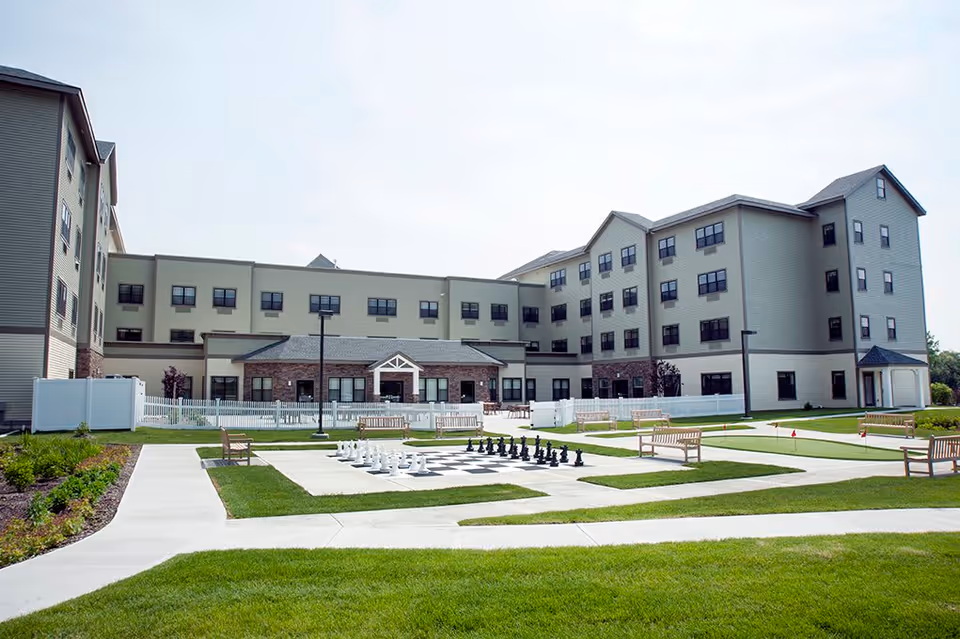 Outdoor courtyard area of Braemar at Wallkill senior living facility featuring a large chessboard with oversized chess pieces, surrounded by benches and pathways, with the multi-story building in the background.