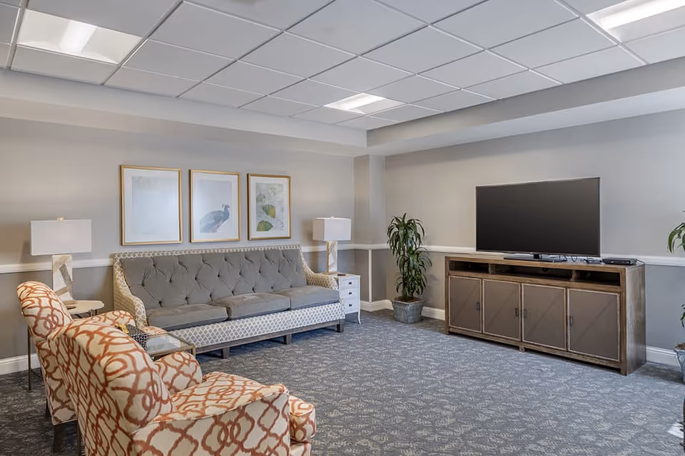 A cozy living room with a gray tufted sofa, two patterned armchairs, two side tables with lamps, a large flat-screen TV on a wooden cabinet, and two potted plants. Three framed pictures hang on the wall above the sofa.