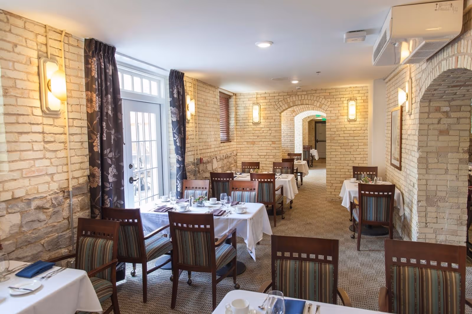 Interior view of a dining room with several tables covered with white tablecloths, set with plates, cups, and silverware. The room features exposed brick walls, arched doorways, patterned curtains, and wooden chairs with striped upholstery. Soft lighting fixtures are mounted on the walls.
