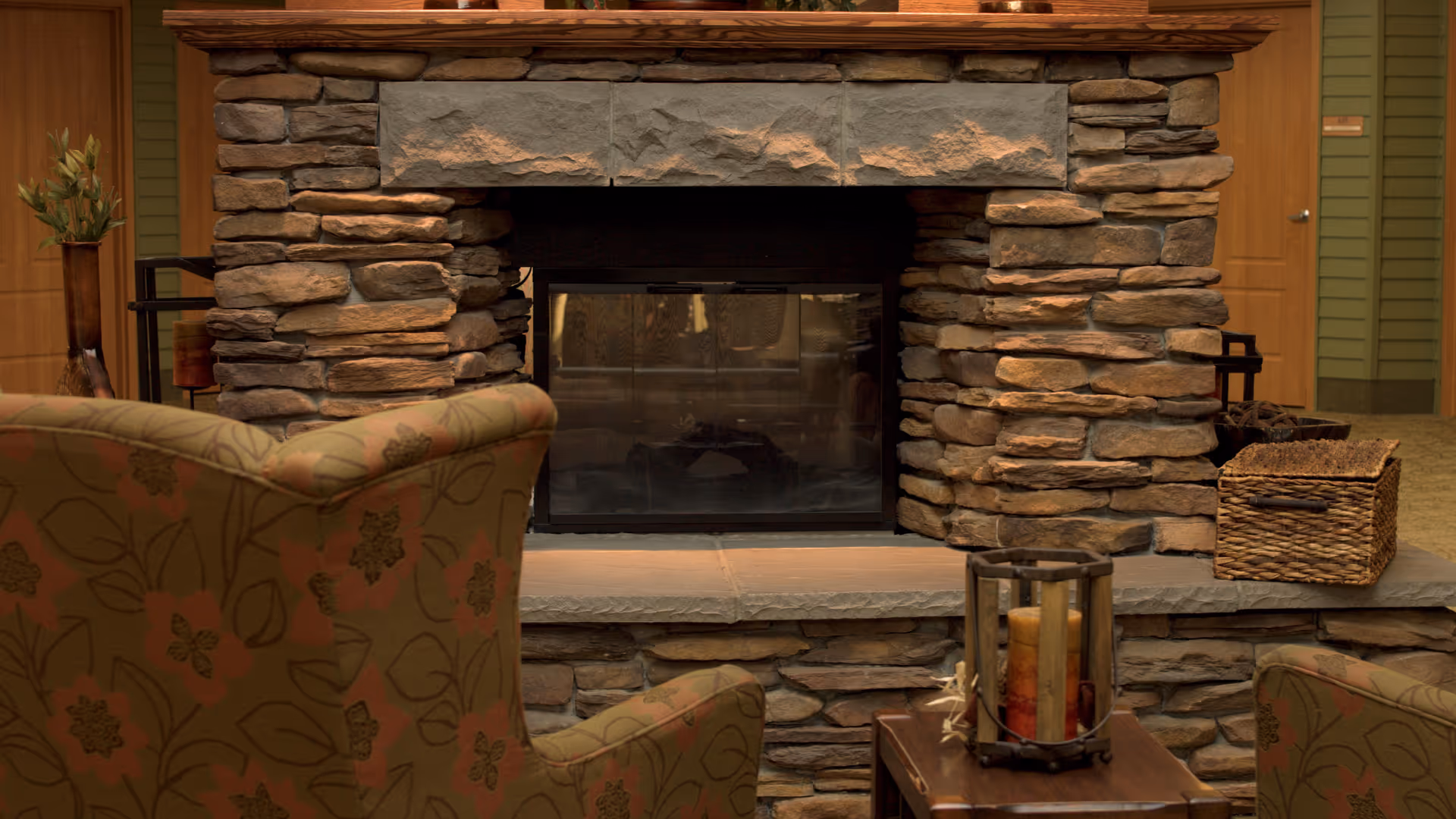Cozy indoor seating area featuring a stone fireplace with a glass front. Two upholstered armchairs with floral patterns face the fireplace. A wooden table in front of the fireplace holds a decorative candle holder with multiple candles. A woven basket and a tall vase with plants are placed on either side of the fireplace. The background includes wooden doors and green paneled walls.