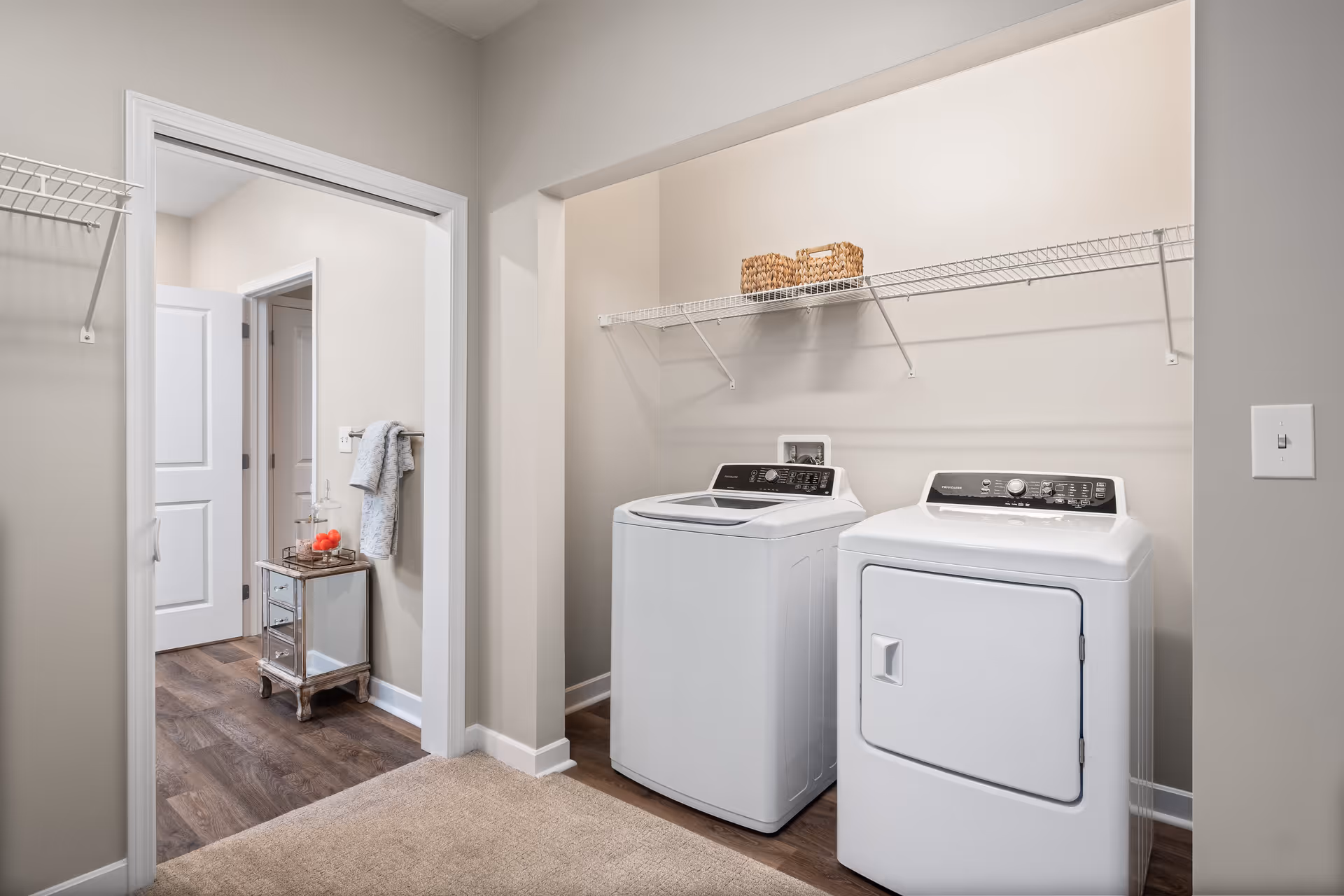 Laundry area with a white top-load washer and matching dryer under a wire shelf, adjacent to a hallway with a small decorative table.
