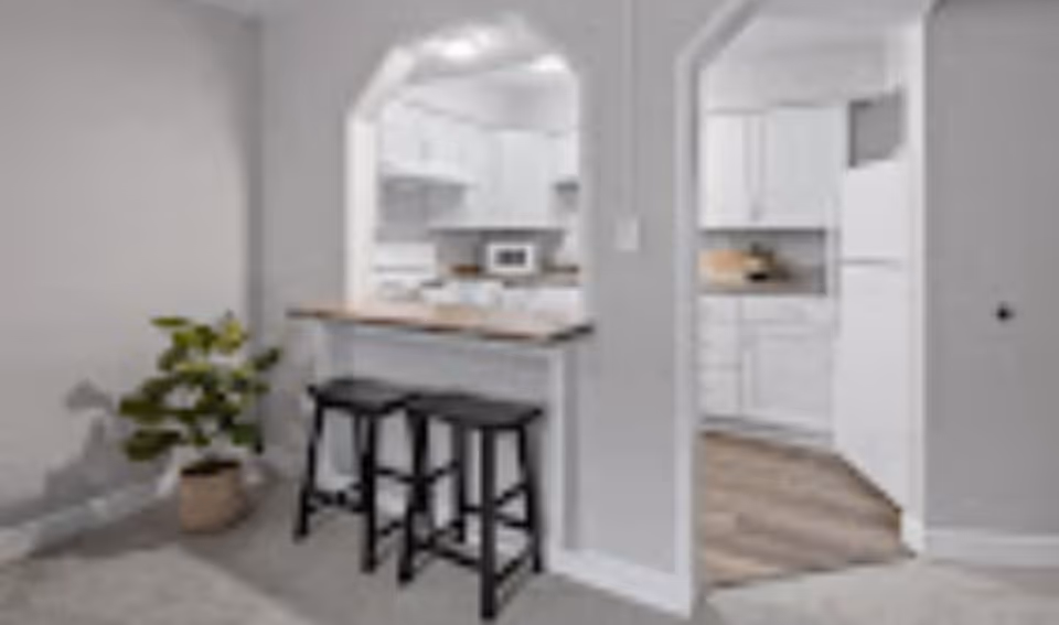 Interior view showing a small breakfast bar with two black stools in front of an open kitchen. The kitchen has white cabinets, a microwave, and a refrigerator. There is a potted plant to the left of the breakfast bar, and the floor transitions from carpet in the foreground to wood flooring in the kitchen area.