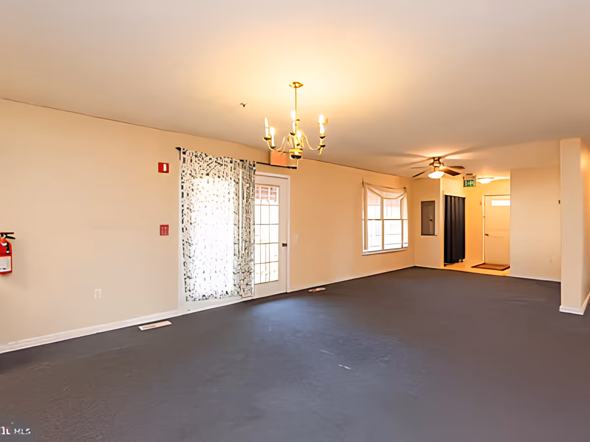 An empty interior room with beige walls and dark flooring. The room features a chandelier hanging from the ceiling, a ceiling fan with light, a window with a curtain, and a glass door with a patterned curtain. There is a fire extinguisher mounted on the wall and an exit sign near a white door at the far end of the room.