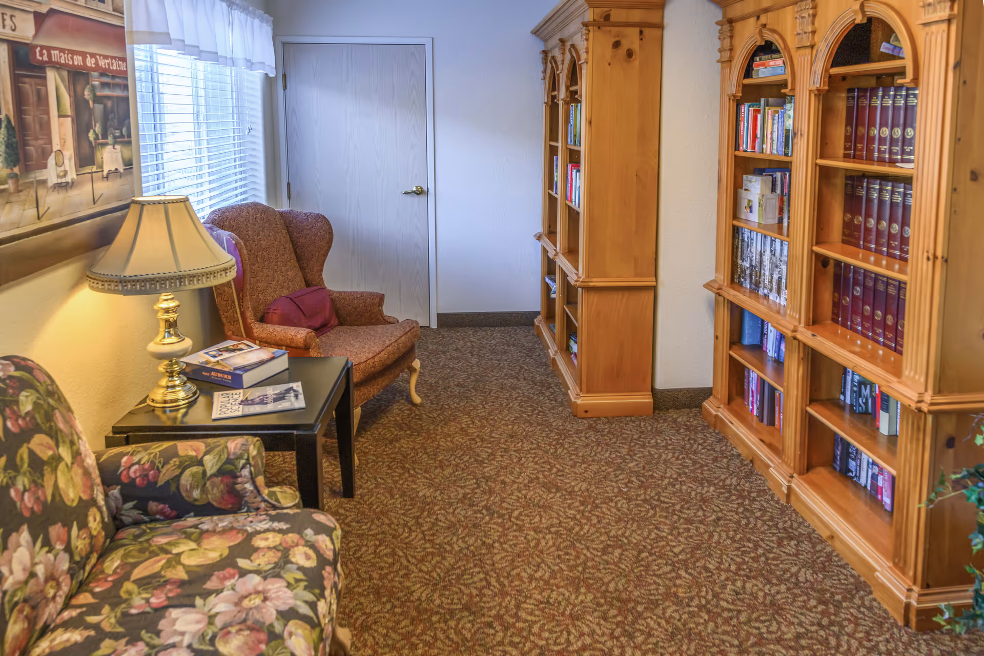 Cozy reading area with floral and wingback chairs, a side table with a lamp, and wooden bookcases along the wall.