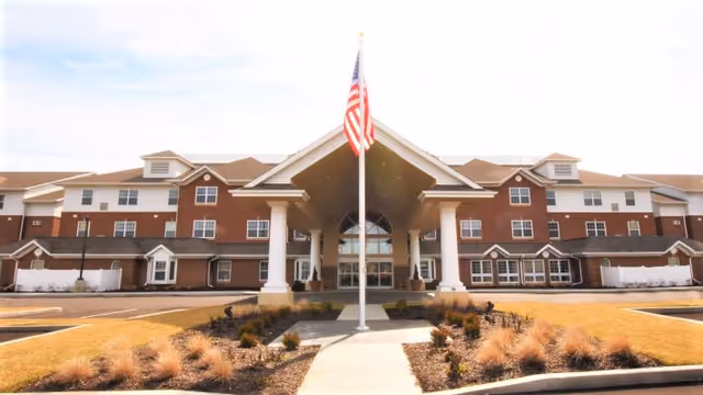 Front entrance of a multi-story brick retirement facility with a covered porte-cochère and an American flag on a flagpole.