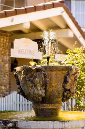 A decorative stone water fountain with water spraying upwards in front of a building entrance with a 'WELCOME' sign and a tiled roof.