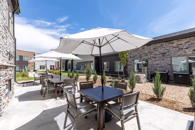 Outdoor patio area at Terra Bluffs with several tables and chairs under large white umbrellas, surrounded by landscaping and buildings with stone and siding exteriors under a blue sky with some clouds.