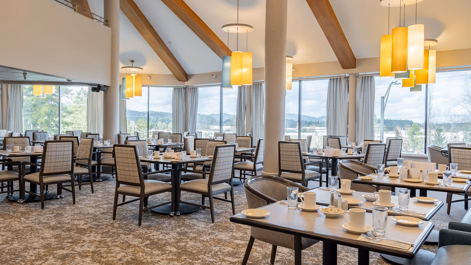 Spacious dining room with multiple tables set with white cups, plates, and glasses. The room features large windows with views of trees and hills outside, beige curtains, carpeted floor, and wooden beams on the ceiling with modern pendant lights hanging down.