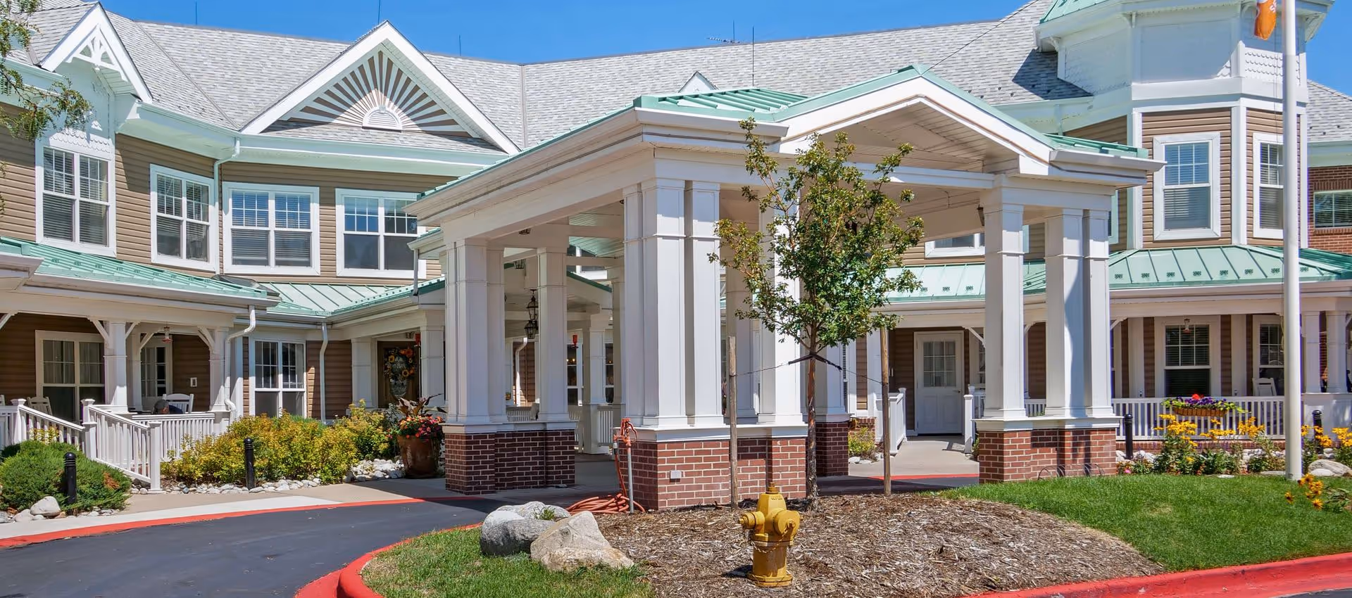 Exterior view of Sunrise at Pinehurst senior living facility showing a covered entrance with white columns and brick bases, a small tree, a yellow fire hydrant, landscaped bushes and flowers, and a building with beige siding, white trim, and green metal roofing under a clear blue sky.