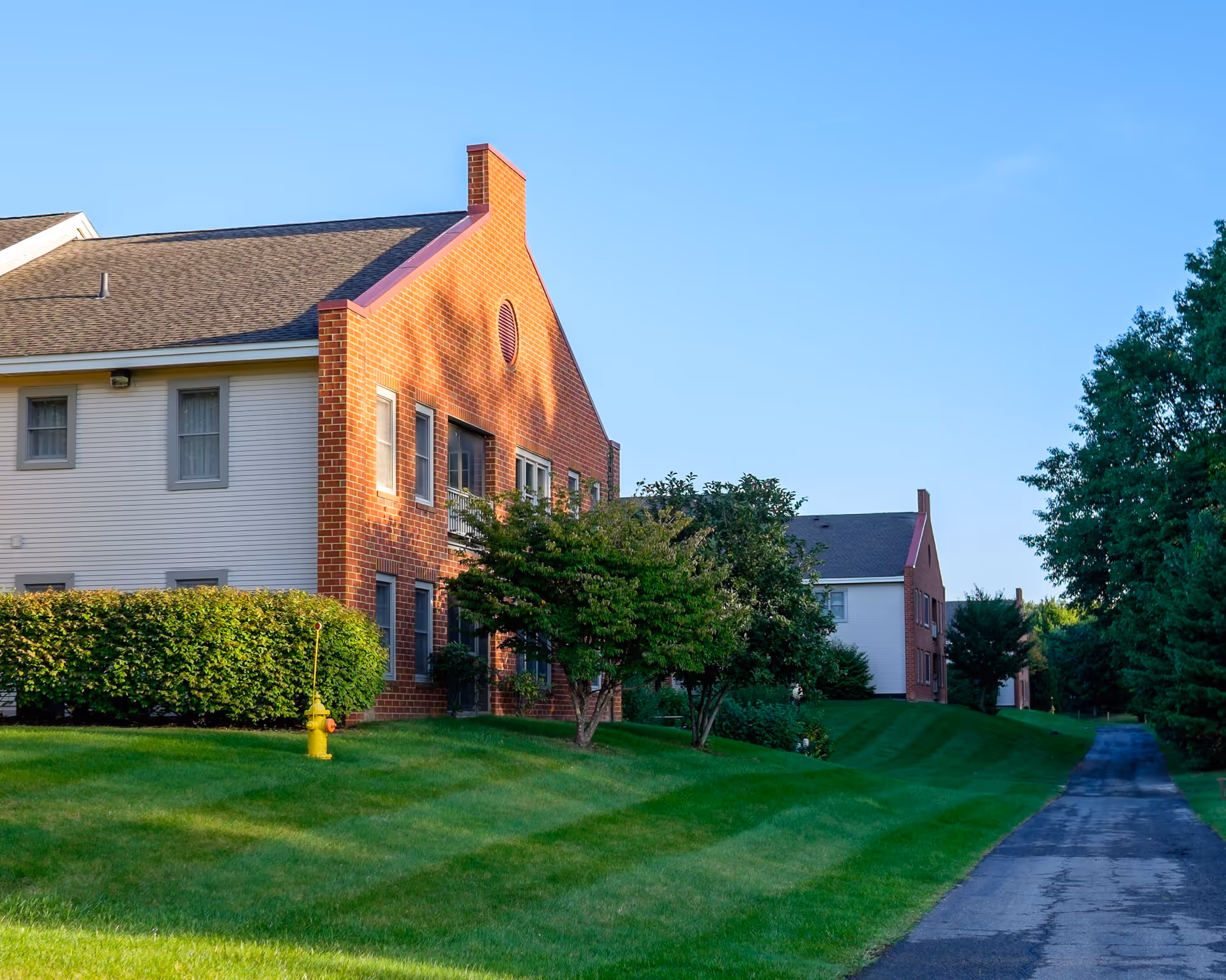 Exterior view of a senior living facility building with brick and siding walls, surrounded by green grass, bushes, and trees under a clear blue sky. A paved pathway runs alongside the building.