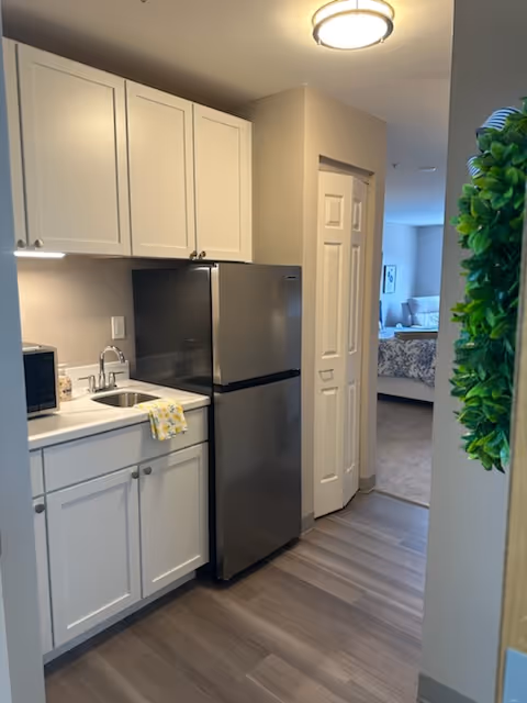 A small kitchen area with white cabinets, a stainless steel refrigerator, a microwave, and a sink with a yellow patterned towel hanging on the cabinet handle. The floor is wood-style laminate, and a doorway leads to a bedroom with a bed and a window in the background. A green leafy decoration is partially visible on the right side of the image.
