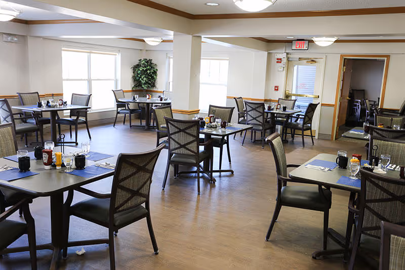 A bright dining room with multiple tables and chairs arranged neatly. Each table has place settings including glasses, napkins, and condiments. Large windows allow natural light to fill the room, and there is a potted plant in the corner near the windows. The floor is wooden, and the walls are painted white with wooden trim. An exit door is visible in the background.