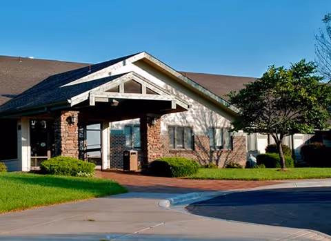 Exterior view of a single-story building with a gabled roof and brick and siding facade, surrounded by green grass, bushes, and a tree under a clear blue sky.