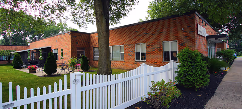 Exterior view of a single-story brick building with several windows, a white picket fence in the foreground, a large tree, and a patio area with tables, chairs, and umbrellas. The surrounding area has green grass, shrubs, and a sidewalk along the right side.
