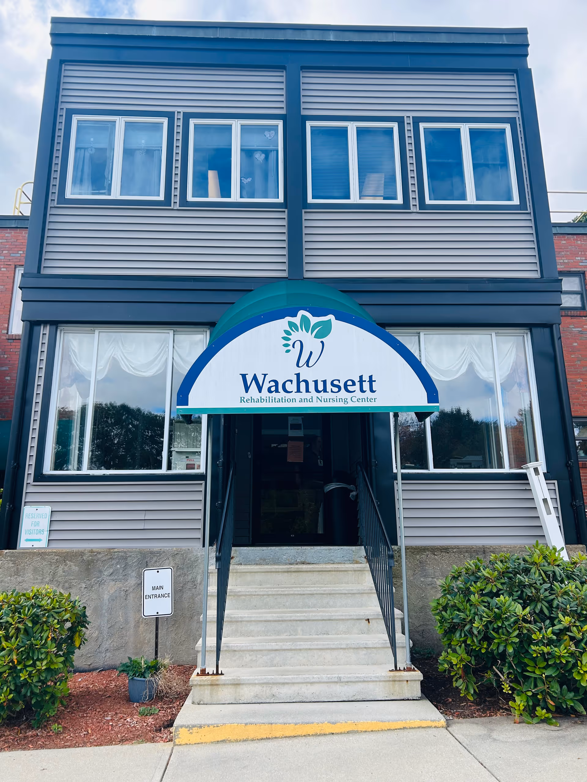 Front entrance of Wachusett Rehabilitation and Nursing Center showing steps leading to a doorway under a branded awning with windows above.