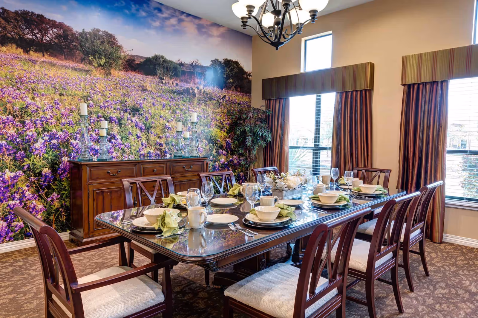 Formal dining room with a long set table and chairs in front of a large floral mural and windows.
