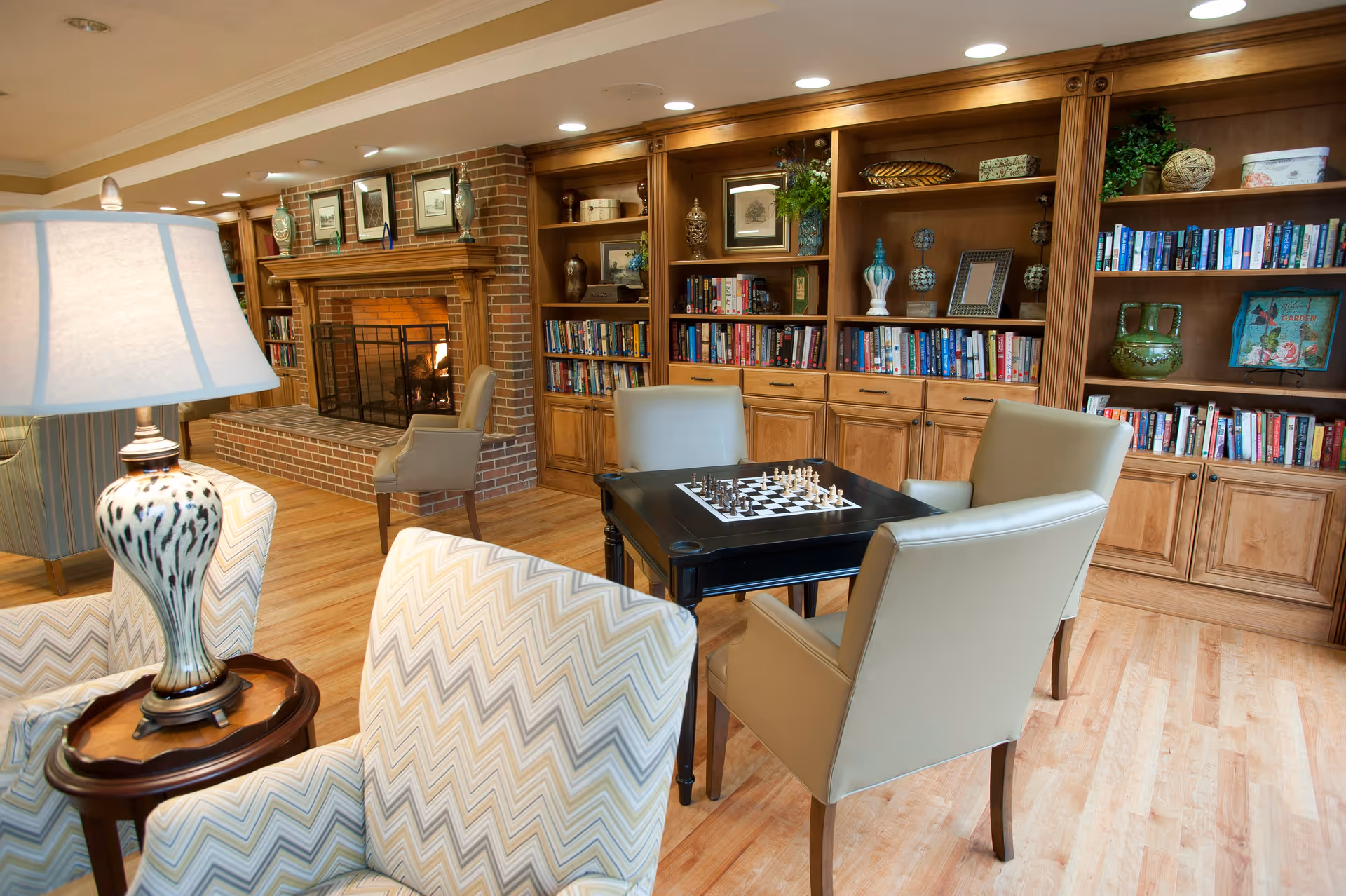 Cozy living room area with a lit brick fireplace, wooden built-in bookshelves filled with books and decorative items, a black table with a chessboard, and several upholstered chairs arranged around the table and near a lamp.