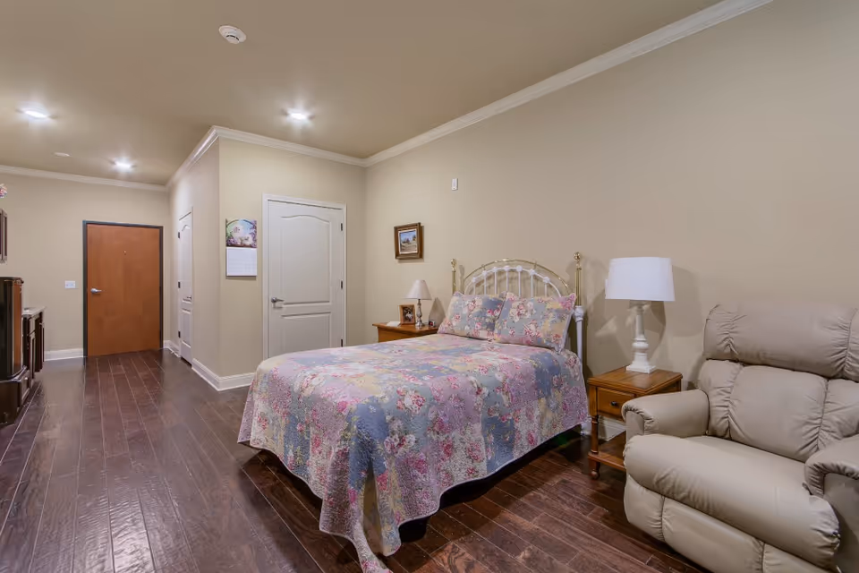 A cozy bedroom with a floral quilted bedspread on a metal bed frame, a beige recliner chair, two wooden nightstands with lamps, and hardwood flooring. The walls are painted beige, and there are two closed doors and a wooden entrance door visible in the background.