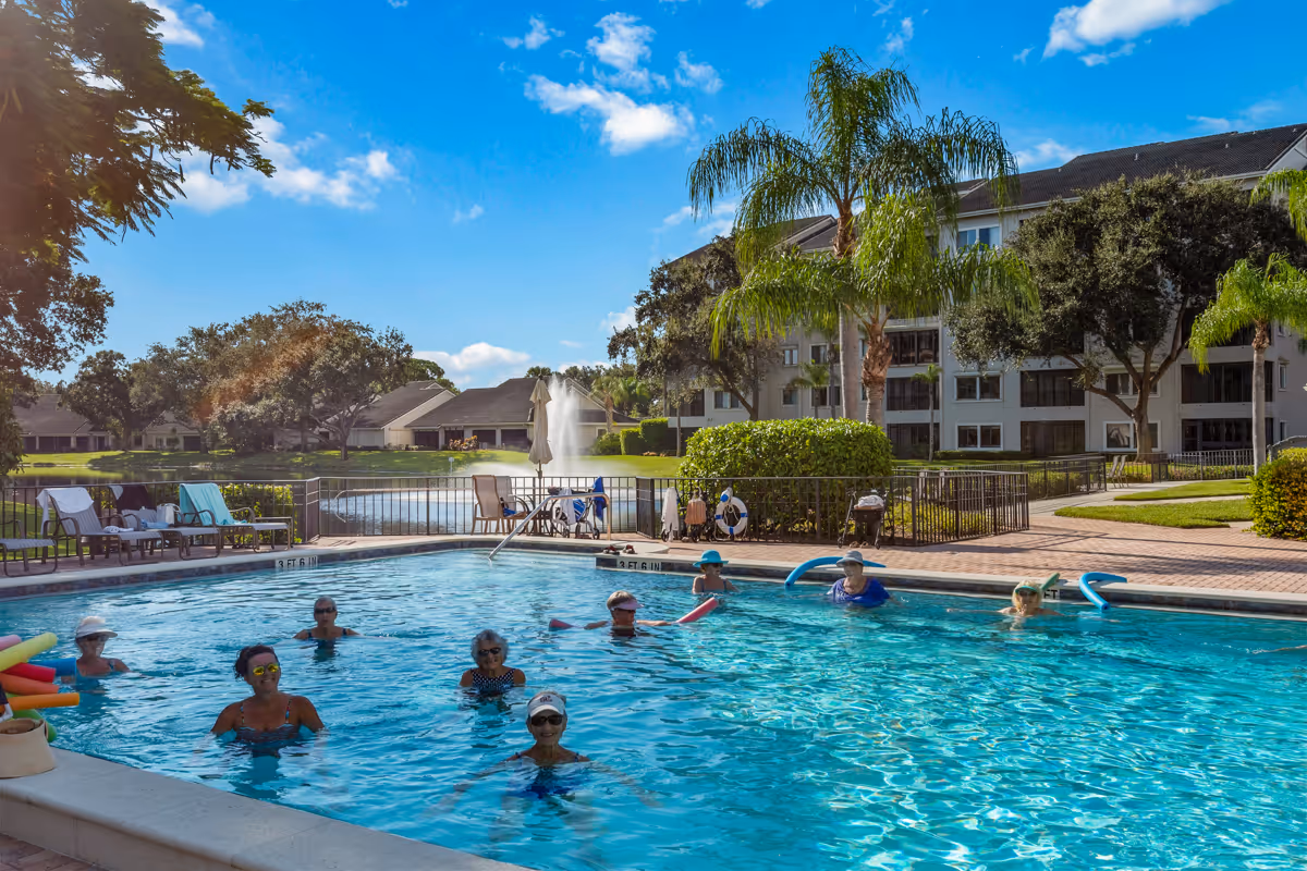 A group of elderly people enjoying a sunny day in an outdoor swimming pool at a senior living facility. The pool area is surrounded by lounge chairs, trees, and a multi-story residential building in the background under a blue sky with some clouds.