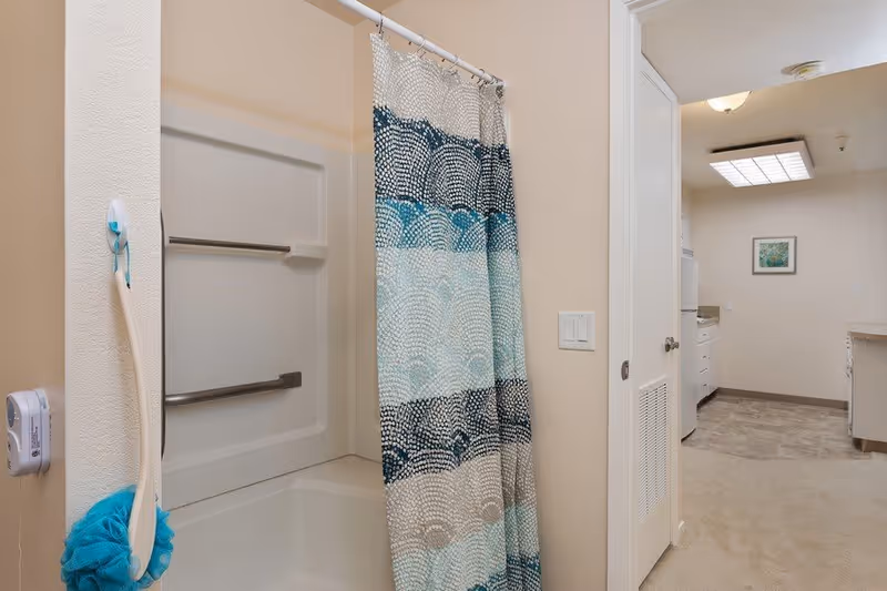 Bathroom with a white bathtub and shower area featuring two metal grab bars. A shower curtain with blue, teal, and white circular patterns is partially drawn. A long-handled bath brush with a blue scrubbing pad hangs on the wall. The bathroom opens into a hallway leading to a laundry area with a stacked washer and dryer, beige walls, and a framed picture on the wall.