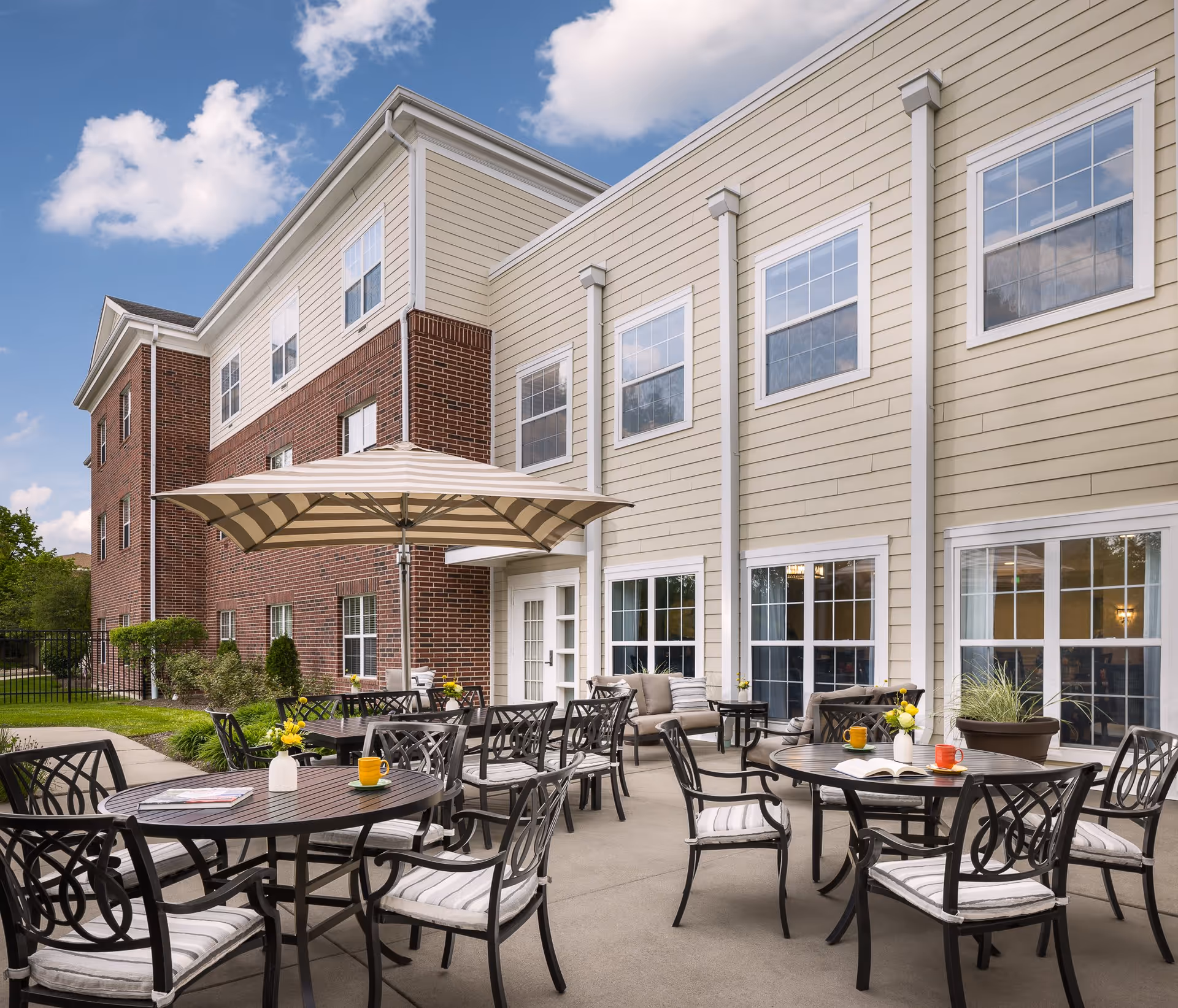 Outdoor patio area at Belmont Village Senior Living Geneva Road with several round tables and chairs, some with striped cushions. A large striped umbrella provides shade over one table. The patio is adjacent to a beige and brick building with multiple windows and glass doors. The sky is partly cloudy.