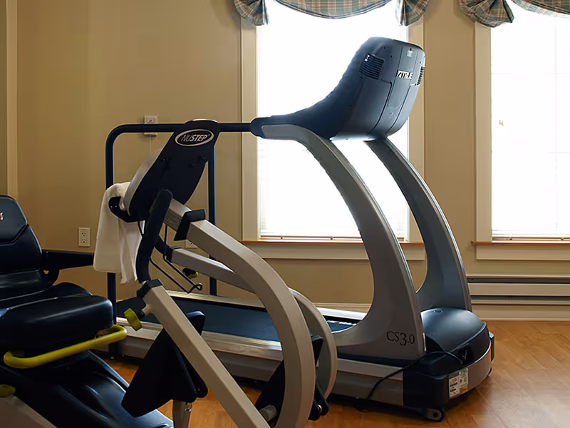 Indoor exercise room with a treadmill and a recumbent exercise bike positioned near windows with blinds and plaid valances.