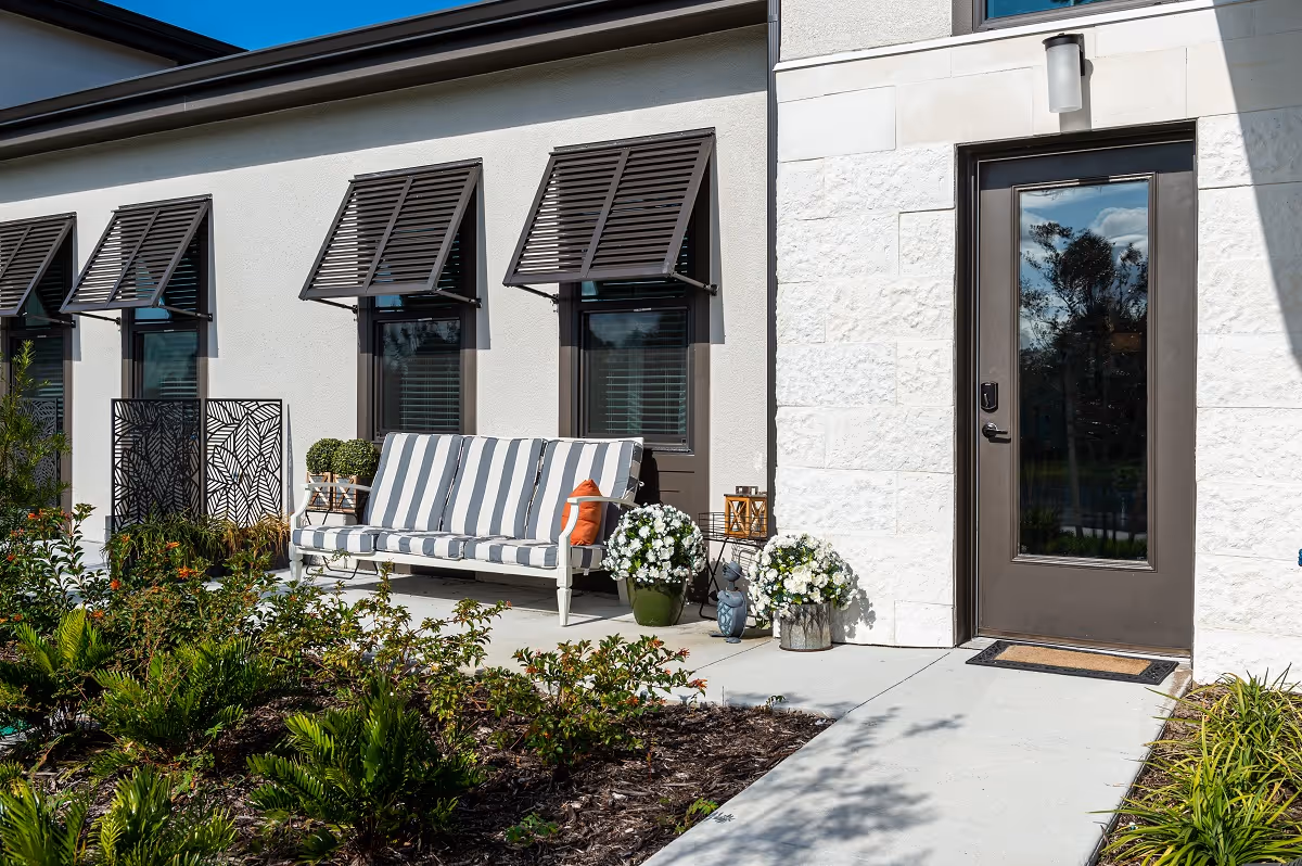 Outdoor patio area with a striped cushioned bench, potted plants, and decorative lanterns next to a door with a glass window. The building exterior features white stone and beige walls with three windows that have angled brown shutters.
