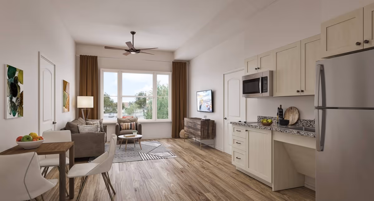A bright and modern living space in a senior living facility featuring a small kitchen area with light wood cabinets, granite countertops, a microwave, and a refrigerator on the right. On the left, there is a dining table with four white chairs and a bowl of fruit. The living area includes a gray sofa, an armchair, a coffee table on a patterned rug, and a wall-mounted TV. Large windows with brown curtains allow natural light to fill the room, and the floor is covered with wood-style flooring.