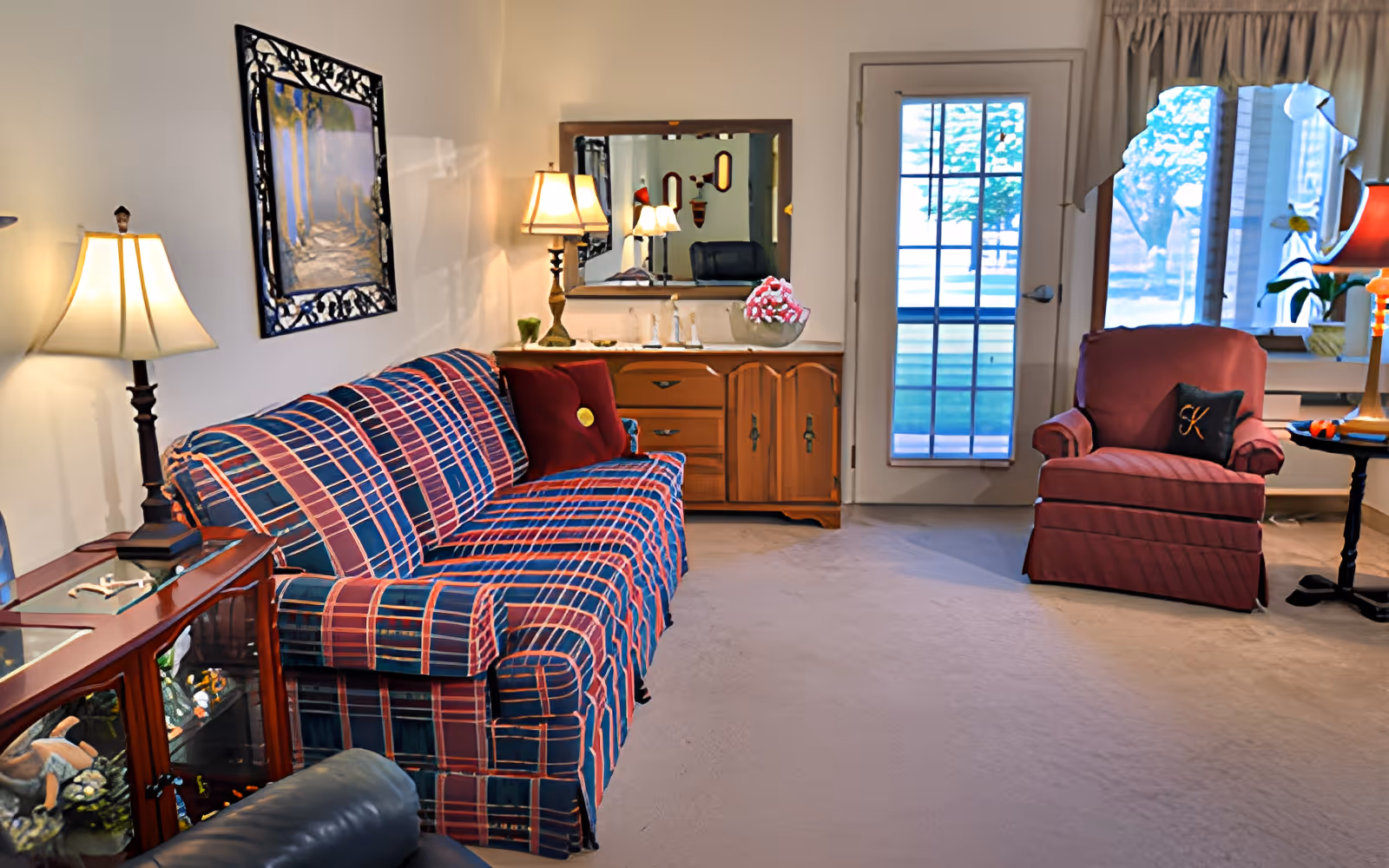 A cozy living room with a plaid-patterned sofa and a red armchair. There are two table lamps on either side of the sofa, a wooden cabinet with a mirror above it, and a glass door leading outside. The room is carpeted and has a window with curtains letting in natural light.