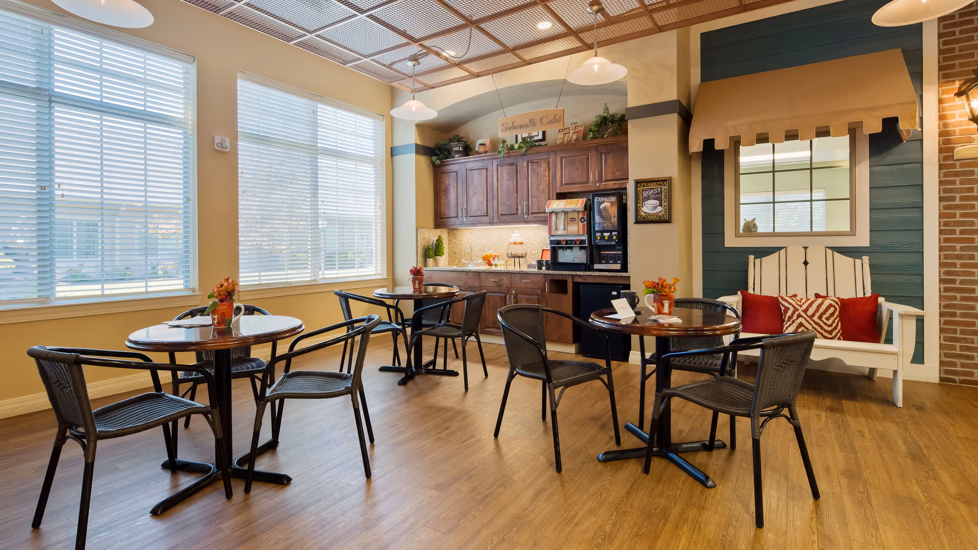 A bright and cozy dining area with round tables and black chairs on a wooden floor. Large windows with white blinds let in natural light. There is a coffee and beverage station with wooden cabinets and a countertop against the wall. A white bench with red and patterned pillows sits under a window with a tan awning, next to a brick accent wall.