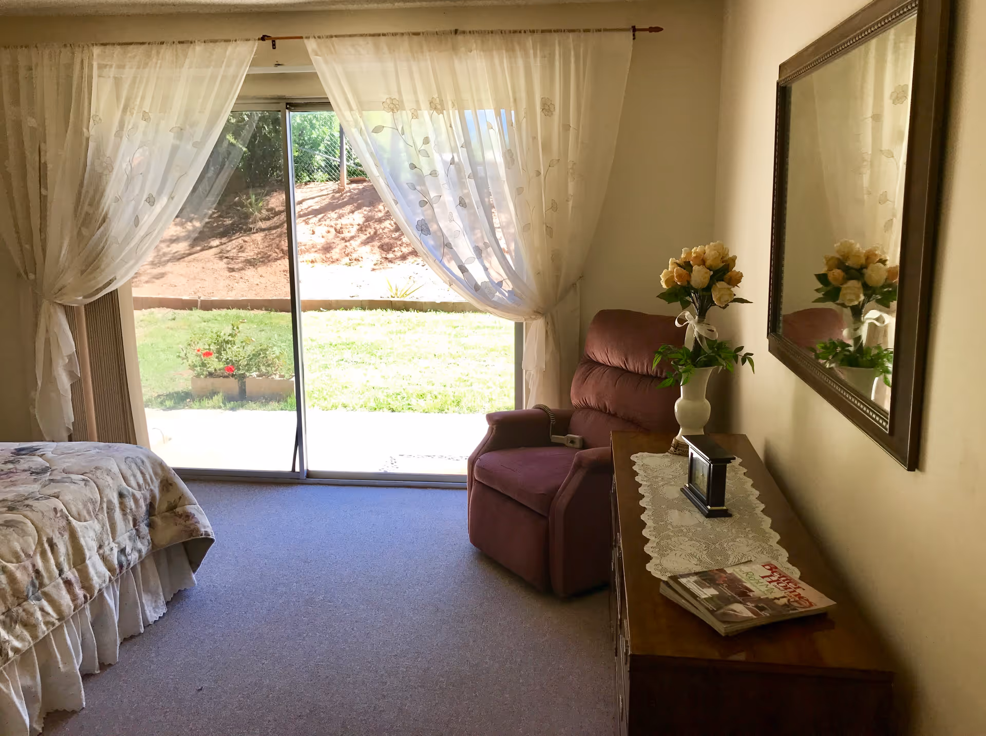 Sunny bedroom with a sliding glass door, recliner, bed corner, and a dresser topped with flowers and a mirror.
