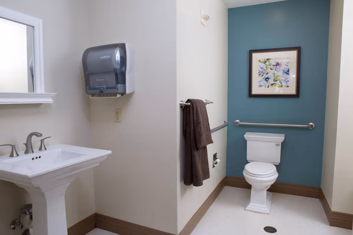 A clean bathroom with a white pedestal sink, a mirror above it, a wall-mounted paper towel dispenser, a toilet with a grab bar on the teal-colored wall behind it, a towel hanging on a wall-mounted towel rack, and a framed floral picture above the toilet.